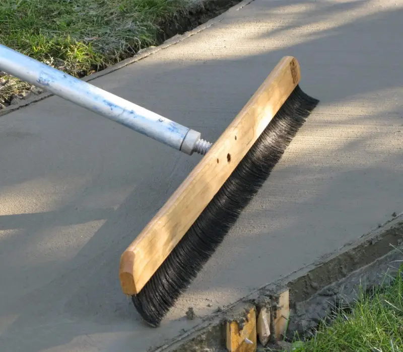 A broom smoothing freshly poured concrete on a sidewalk.
